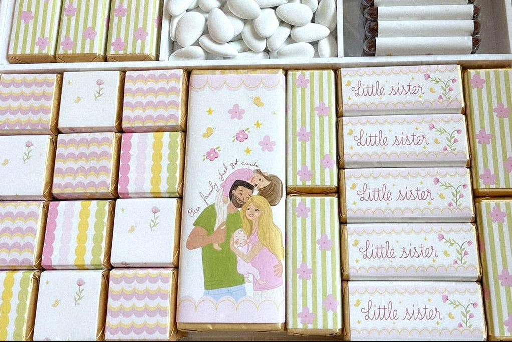 A boxed assortment of pastel-themed chocolates and candy bars arranged in a square tray. The set includes striped and floral wrapped bars, white chocolate-covered pieces, candy-coated chocolates, and a central card with an illustration of a girl in a pink hat.
